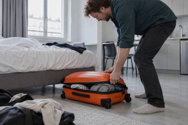 Fiktiv rejsende pakker en orange kuffert ved en seng i en feriebolig; ikke en konkret Airbnb-bolig, vært eller dokumentation af konflikten. AI-genereret, fotorealistisk temabillede; viser ikke den konkrete begivenhed.
