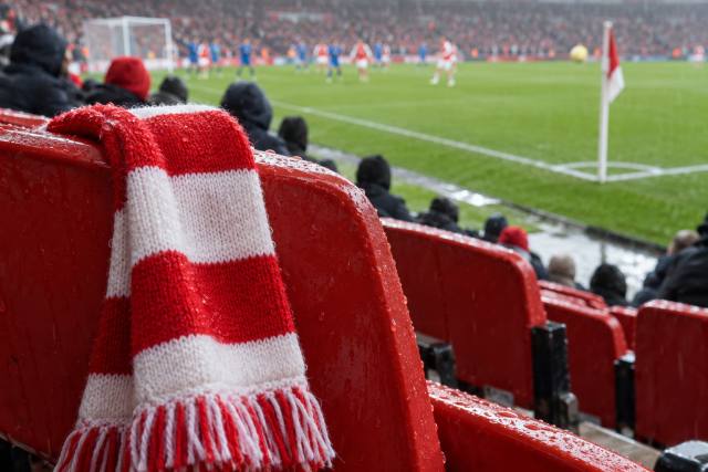 Rødt og hvidt halstørklæde på et regnvådt rødt stadionsæde ved en fiktiv fodboldbane; ikke Emirates Stadium eller et foto fra Arsenal–Everton. AI-genereret, fotorealistisk temabillede; viser ikke den konkrete begivenhed.