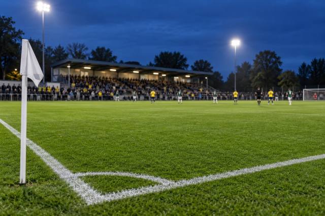 AI-genereret, fotorealistisk temabillede af en fiktiv fodboldbane med hvidt hjørneflag og tilskuere under projektører; ikke Gladsaxe Stadion, AB–Brøndby eller dokumentation af den konkrete kamp eller tilskuerrekord.
