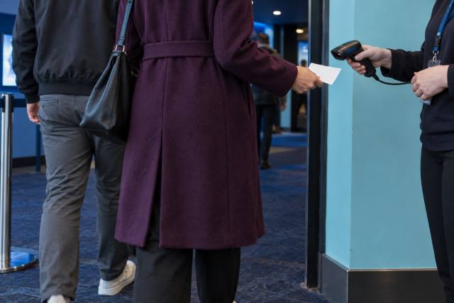 Fiktive voksne biografgæster ved billetkontrol i en cyanfarvet foyer; ikke en konkret Clayface-forestilling eller et billede fra filmen. AI-genereret, fotorealistisk temabillede; viser ikke den konkrete begivenhed.