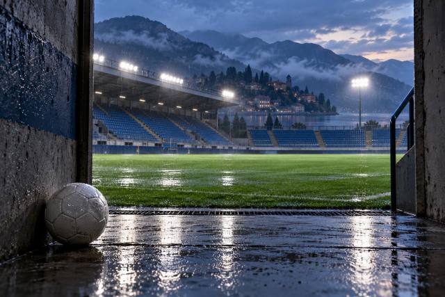 Fiktivt fodboldstadion i et norditaliensk bjerglandskab set fra en tunnel i regnvejr; ikke Comos stadion eller et foto fra kampen. AI-genereret, fotorealistisk temabillede; viser ikke den konkrete begivenhed.